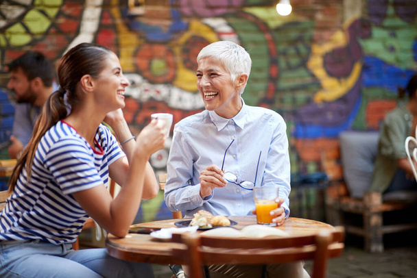 Dos amigas felices de diferentes generaciones que se divierten mientras toman una copa en un ambiente alegre en el bar. Ocio, bar, amistad, al aire libre - Foto, Imagen