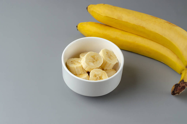 Two bananas and slices in a white plate on a gray background. Trending color of year 2021 Illuminating and Ultimate gray. Side view minimal still life. - Photo, image