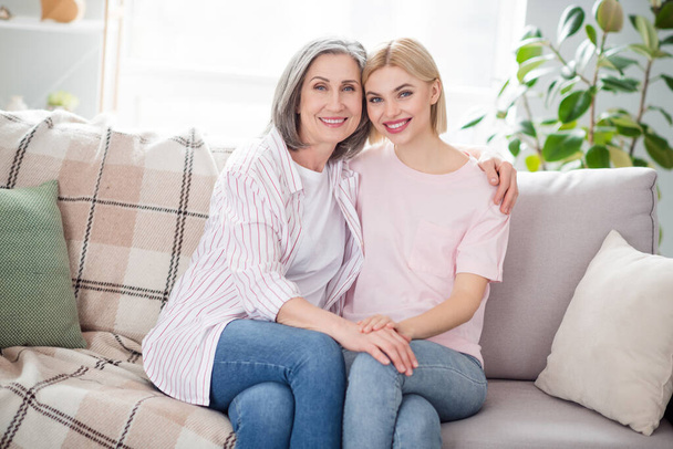 Photo of two pretty positive ladies sit on couch hugging touch hand toothy smile look camera have good mood indoors - Foto, Bild