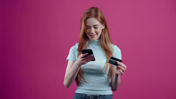 The happy young woman with the phone and the card in her hand is shopping online, in a blue casual t-shirt, isolated on a pink background. The concept of peoples lifestyle - 映像、動画