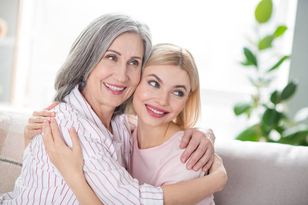 Portrait of two charming cheerful ladies sit on sofa hugging each other look far away beaming smile indoors - Photo, Image