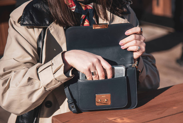 Una mujer en un impermeable ligero pone su teléfono en su bolso, sentado en un café abierto en un día soleado. - Foto, imagen