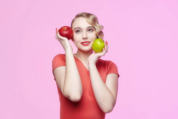 attractive young blonde with a retro hairstyle holds a red and green apple in front of her face - Foto, imagen