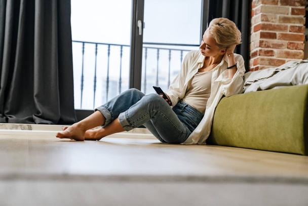 Жінка розслабляється, спираючись на диван, використовуючи смартфон вдома
 - Фото, зображення