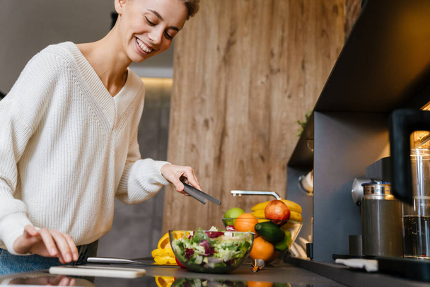 Beautiful smiling young woman using mobile phone while making healthy salad in the kitchen - 写真・画像