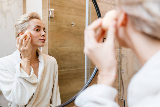 Attractive young woman wearing bathrobe applying makeup with sponge while looking in the mirror in bathroom - Photo, image