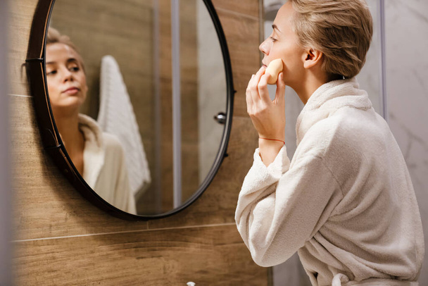 Attractive young woman wearing bathrobe applying makeup with sponge while looking in the mirror in bathroom - Zdjęcie, obraz