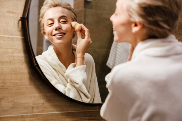 Attractive young woman wearing bathrobe applying makeup with sponge while looking in the mirror in bathroom - Foto, immagini