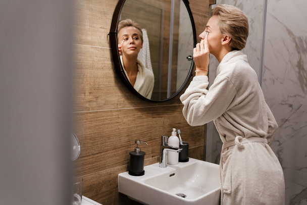 Woman in bathroom cleaning her face with cotton bud, looking in the mirror - Photo, image