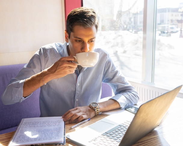 a young businessman looks into a laptop and drinks cappuccino coffee, a notebook with his notes lies next to him, he is in a cafe - Foto, immagini