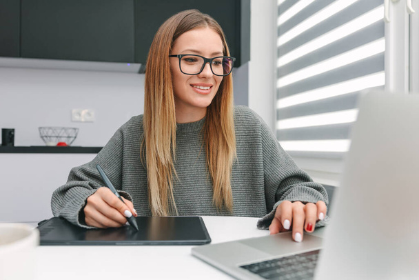 Smiling female designer with glasses working on graphic tablet on laptop at home - Фото, изображение
