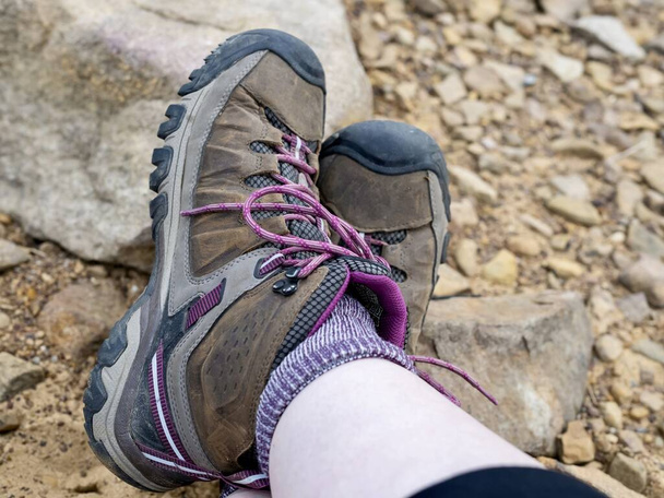 Imagen de cerca de un par de botas de senderismo marrones y rosados con calcetines de lana púrpura con un fondo arenoso rocoso, listo para la aventura y los senderos. - Foto, imagen
