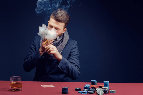 Stylish bearded Man in suit and scarf playing in dark casino, smoking cigar, drink whiskey - Fotografie, Obrázek