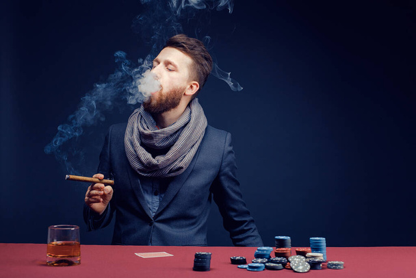 Stylish bearded Man in suit and scarf playing in dark casino, smoking cigar, drink whiskey - Photo, Image