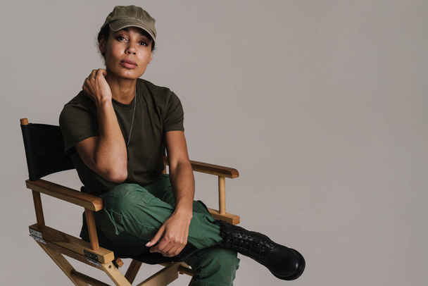 Confident african american soldier woman looking at camera while sitting on chair isolated over grey wall - Фото, изображение
