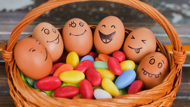Stos jaj z ręcznie narysowanymi twarzami na słomkowym koszu z kolorowymi cukierkami, przygotowanie wielkanocne, świąteczne koncepcje nastroju - Zdjęcie, obraz