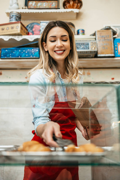 Beautiful young female seller working in a sweet food house or bakery. She is selling and packing delicious sweets. - 写真・画像