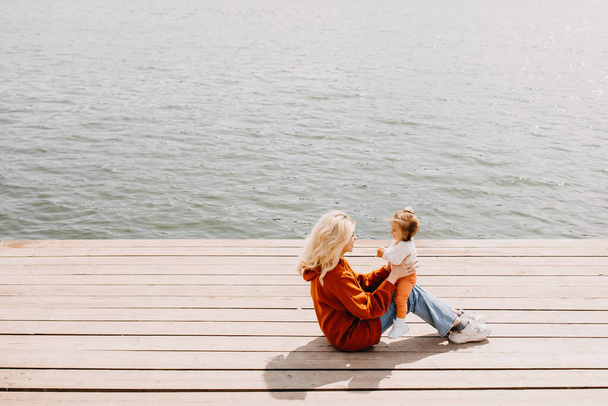 Молода мати грає зі своєю дочкою на відкритому повітрі, сидячи на дерев'яному пірсі біля озера
. - Фото, зображення