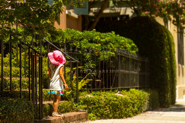 A cute little caucasian girl with tanned skin is standing on the stairs at the entrance of her house on a sunny summer day. She wears short skirt and sleeveless blouse as well as a wide brim solar hat - 写真・画像