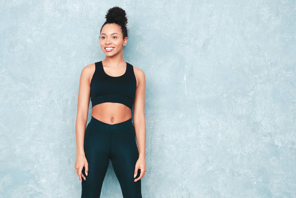 Fitness smiling black woman in sports clothing with afro curls hairstyle.She wearing sportswear. Young beautiful model with perfect tanned body.Female posing in studio near gray wall  - 写真・画像