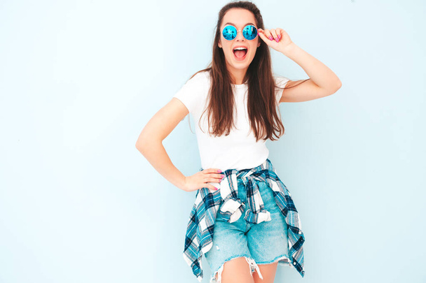 Young beautiful smiling female in trendy summer hipster clothes. Sexy carefree woman posing near light blue wall in studio. Positive model having fun indoors. Cheerful and happy in sunglasses - Zdjęcie, obraz