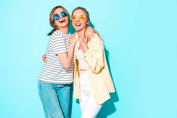 Two young beautiful blond smiling hipster female in trendy summer clothes. Sexy carefree women posing near blue wall in studio. Trendy and positive models having fun in sunglasses - Fotoğraf, Görsel