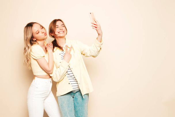 Two young beautiful blond smiling hipster female in trendy summer clothes. Sexy carefree women posing in studio. Trendy and positive models having fun.Taking selfie photos - Zdjęcie, obraz