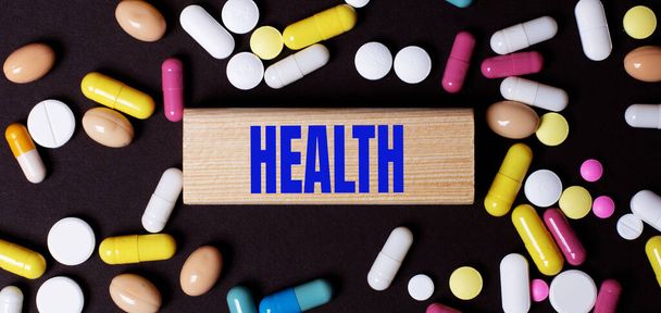 On a dark background, multi-colored pills and the word HEALTH on a wooden block. Medical concept - Foto, afbeelding
