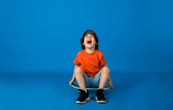 cheerful little boy with brown hair in a T-shirt and denim shorts sits on a skateboard on a blue background with space for text. Street sports - Photo, Image