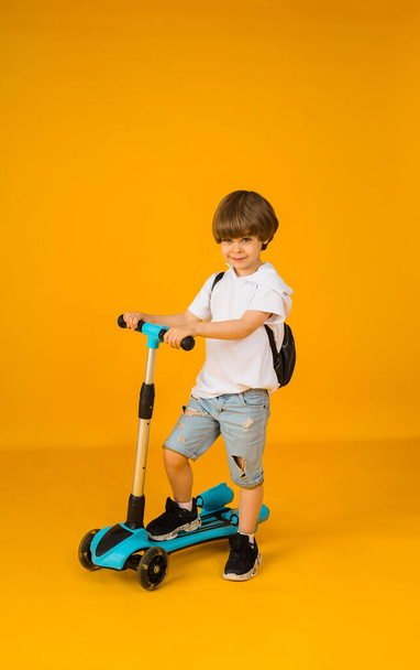 little boy in a white T-shirt and denim shorts rides a scooter on a yellow background with space for text. Street sports - Foto, Imagem