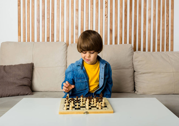 zamyślony mały biały chłopiec z brązowymi włosami siedzi na kanapie i gra w szachy. Rozwój dziecka - Zdjęcie, obraz