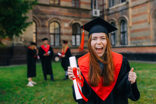 笑顔、喜び、赤髪の女の子、叫んで、彼女の手の中に卒業証書を振って。卒業生のガウンとピークキャップ. - 写真・画像