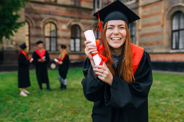 Χαρούμενη απόφοιτη με ακαδημαϊκό φόρεμα. Δείχνει έναν πάπυρο με κόκκινη κορδέλα. Απολαύστε την αποφοίτηση από το πανεπιστήμιο. - Φωτογραφία, εικόνα