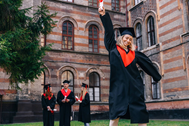 Boldog mosolygó diplomás lány fekete köpenyben felemeli a kezét egy diplomával a kezében. Diák pózol az egyetemi udvaron az osztálytársak hátterével szemben. Sikerkoncepció. - Fotó, kép