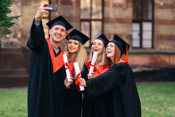 concepto de educación, graduación, tecnología y personas - grupo de estudiantes felices en tablas de mortero y vestidos de soltero con diplomas tomando selfie al aire libre. - Foto, Imagen