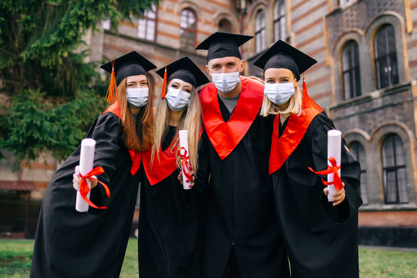 Un graduado con máscaras médicas en la cara. Estudiantes felices con diplomas en un vestido académico y gorra de graduación, un grupo con un certificado de educación, están de pie cerca de la universidad. - Foto, Imagen