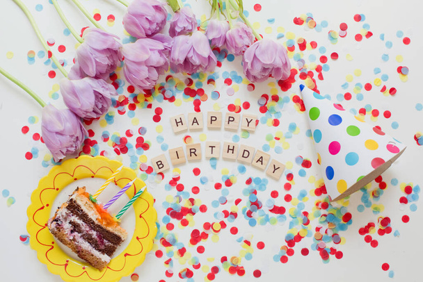 Születésnapi háttér ízletes torta egy tányéron, papír party kalap, színes konfetti és fa felirat betűk - Fotó, kép