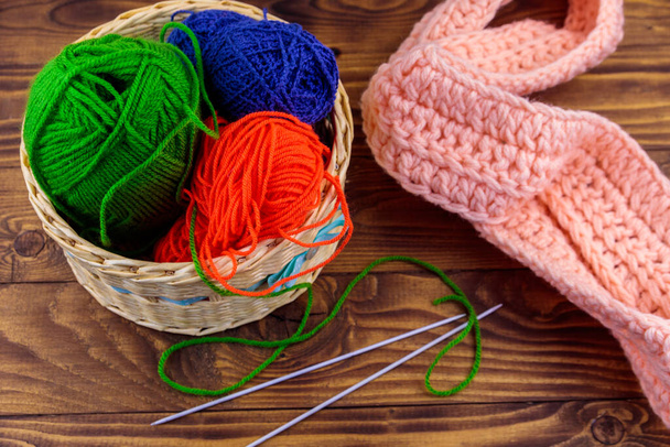 Плетеная корзина с красочными вязальными нитями, вязальными спицами и вязаным шарфом на деревянном столе - Фото, изображение