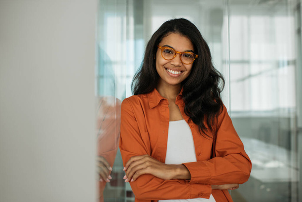 現代のオフィスに立ってカメラを見てスタイリッシュな眼鏡をかけて笑顔アフリカ系アメリカ人のビジネス女性の肖像画。成功したビジネスとキャリアの概念  - 写真・画像
