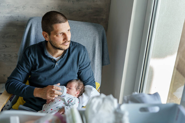 Kaukázusi férfi férfi apa áll az ablaknál otthon gazdaság és palack etetés tej az újszülött lánya vagy fia a nap apák napja szülőség valós emberek koncepció másolás tér - Fotó, kép