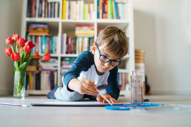 Edessä näkymä nelivuotias valkoihoinen poika leikkii lattialla kotona pieni lapsi käyttää sivellintä maalatessaan akvarelleja kehittää taiteen taitoja kasvaa käsite vapaa-ajan ja luovan koulutuksen - Valokuva, kuva