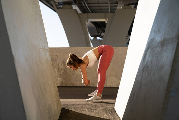 Νεαρή αθλήτρια προετοιμάζεται για προπόνηση, τεντώνει τους μυς, σκύβει μπροστά κάτω από τη γέφυρα. Fit σπορ κορίτσι ζεσταθεί πριν από μαραθώνιο τρέξιμο. Γυμναστική, σωματική και ψυχική υγεία και τρόπος ζωής - Φωτογραφία, εικόνα