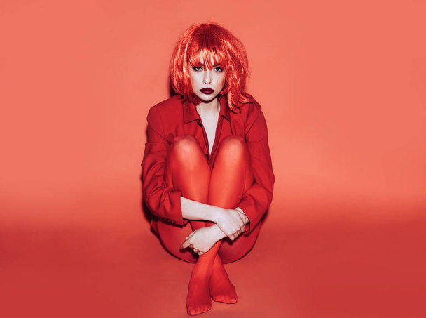 Woman in red wig, tights and jacket sitting on floor with legs crossed. Teenage girl with offended look isolated on red background. - Foto, imagen