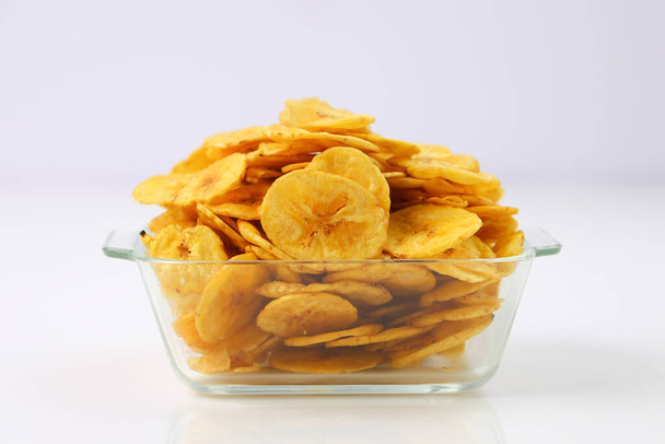 Suszone chipsy bananowe lub gofry bananowe, pięknie ułożone w szklanej misce lub zastawie stołowym z białym, teksturowanym tłem, izolowane. - Zdjęcie, obraz