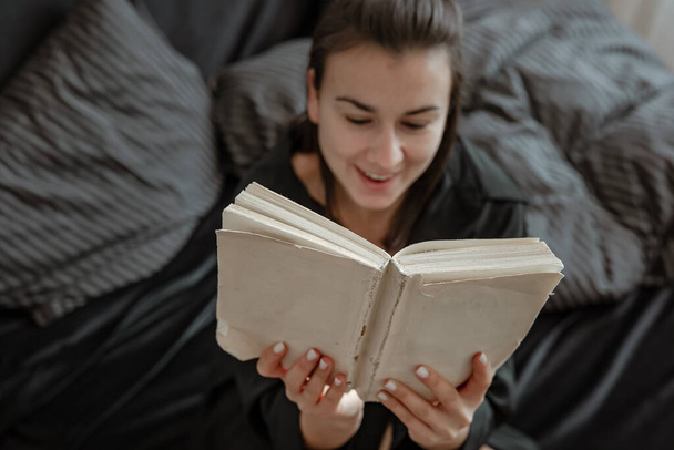 Ελκυστική νεαρή γυναίκα με πιτζάμες χαλαρώνει στο κρεβάτι διαβάζοντας ένα βιβλίο. - Φωτογραφία, εικόνα