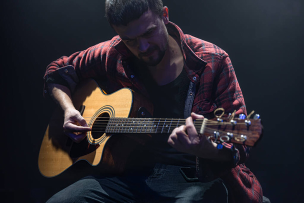 Muzyk gra na gitarze siedząc w ciemnym pokoju. - Zdjęcie, obraz