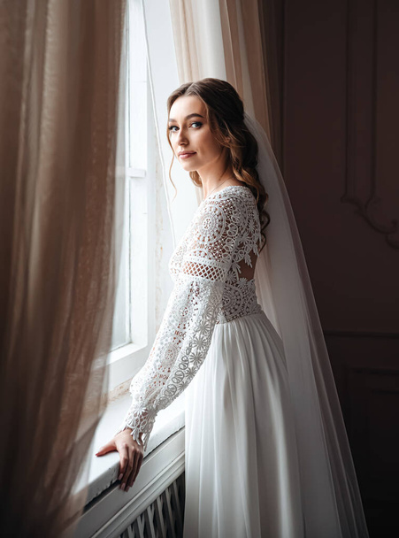 Eine junge Braut in einem wunderschönen Boho-Spitzenkleid steht am Fenster. Hochzeits-Fotosession, Brautmorgen. - Foto, Bild