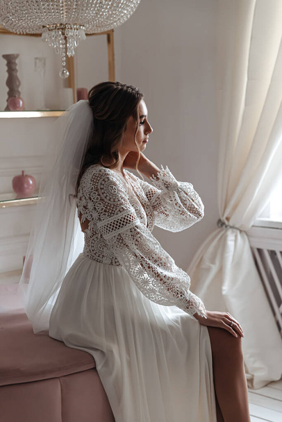Menyasszony egy esküvői ruha boho stílusban, és egy fátyol pózol ül egy hangulatos szobában. Esküvői fotózás, menyasszony reggele. - Fotó, kép