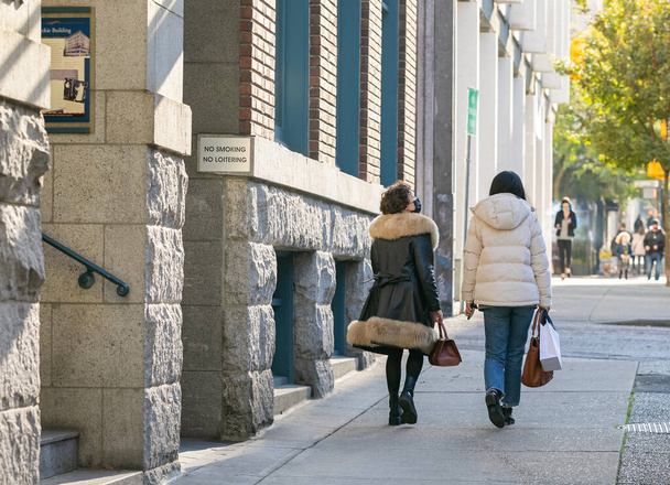 Zwei junge Frauen in lässiger, stylischer Kleidung gehen mit Einkaufstüten auf die Straße. Street View, Reisefoto, selektiver Fokus. 1.November 2020-Vancouver, BC, Kanada - Foto, Bild