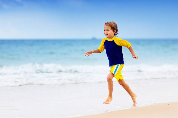 Παιδιά που παίζουν σε τροπική παραλία. Τα παιδιά κολυμπούν και παίζουν στη θάλασσα στις καλοκαιρινές οικογενειακές διακοπές. Άμμος και νερό διασκέδαση, προστασία από τον ήλιο. Μικρό παιδί τρέχει και πηδάει στην ακτή του ωκεανού. - Φωτογραφία, εικόνα